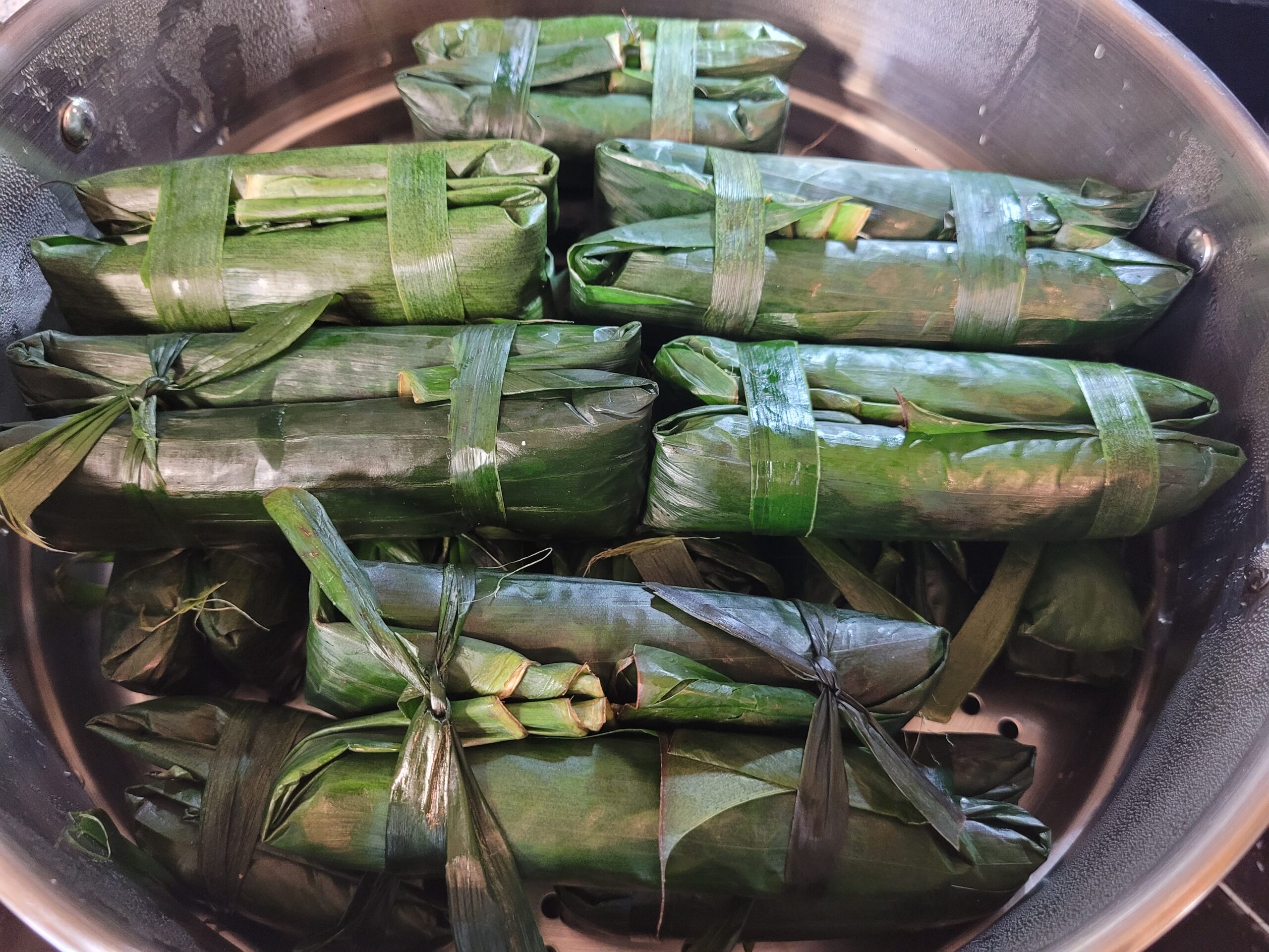 Steamed Filipino suman wrapped in banana leaves and tied with leaf strips, arranged in a stainless-steel steamer basket during cooking.